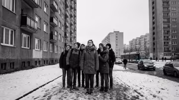 Širokoúhlý záběr z nízkého úhlu zachycuje skupinku teenagerů na zasněžené betonové cestě mezi dvěma holskými panelákovými bloky.